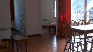 A spacious living area with a dining table and chairs, a large window with a view of buildings, and a door leading to another room. - Les Hauts Plateaux (Ax-les-Thermes)