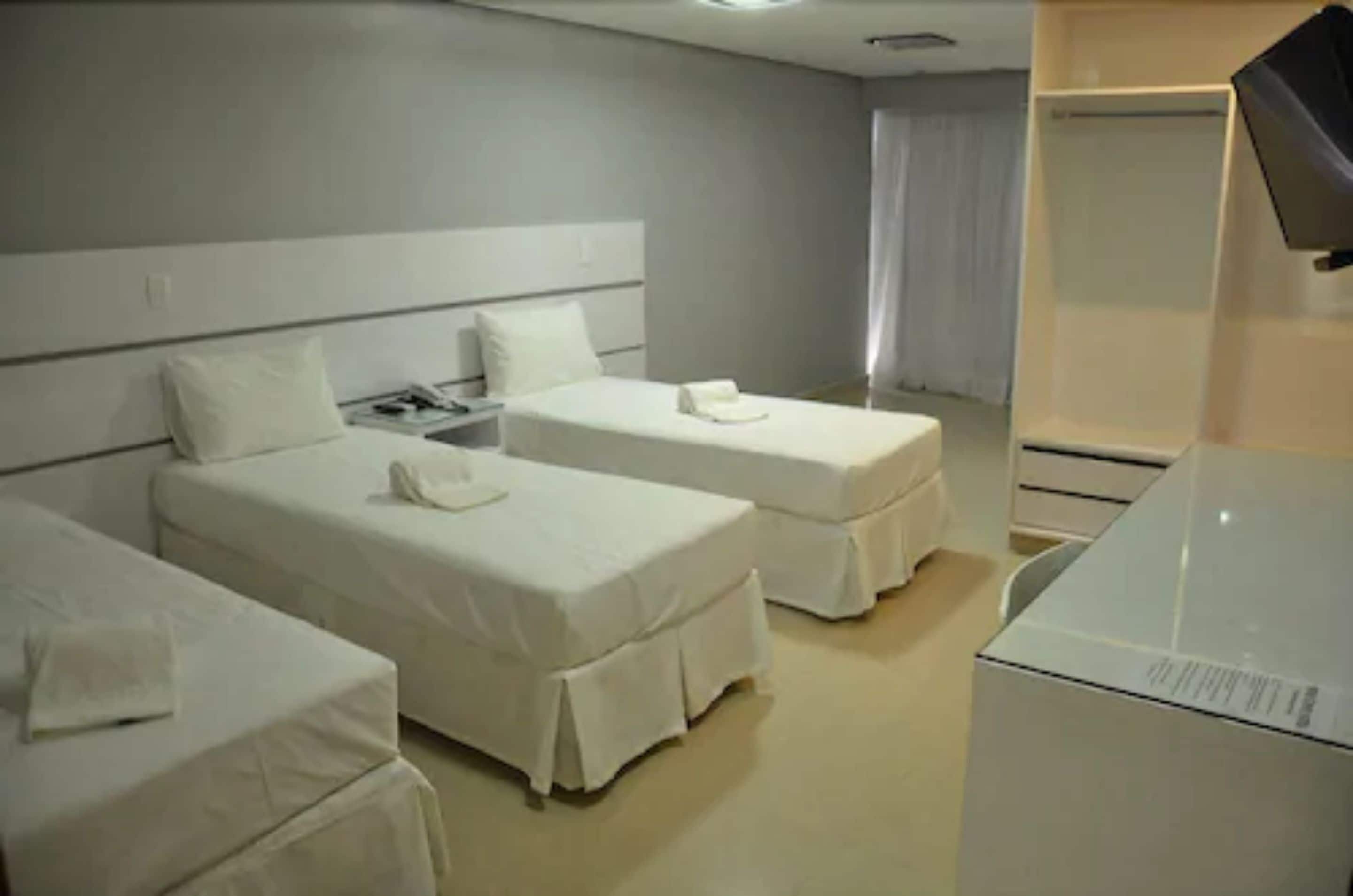A hotel room with two single beds, a bedside table with a lamp, a small table with a phone, and a large white appliance, likely a refrigerator or washer.