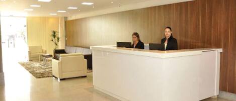 A reception area with a white reception desk, two receptionists, a sofa, and a wooden wall.