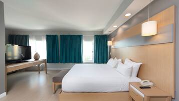 Una habitación de hotel moderna con una cama grande, un televisor sobre un mueble de madera, una mesita de noche con un teléfono y cortinas color turquesa.