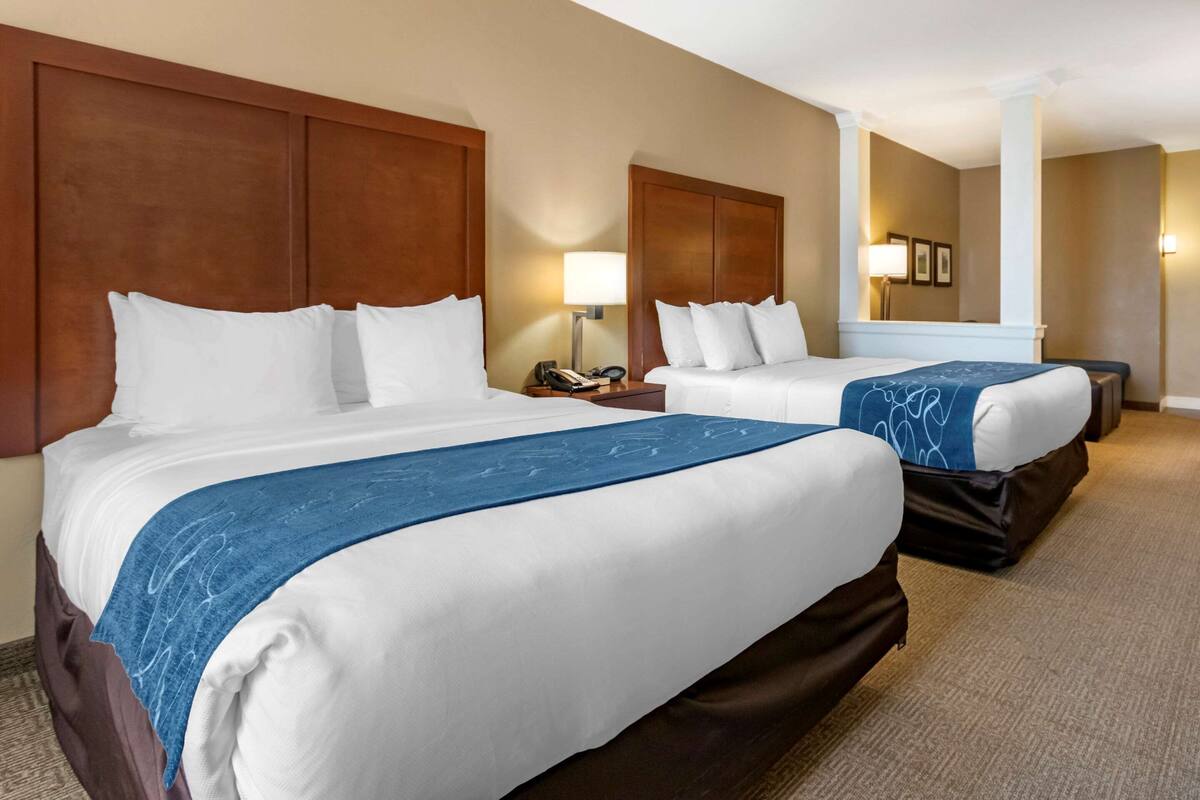 A hotel room with two beds, each with white linens and a blue accent. There are bedside tables with lamps and a phone. The room has a wooden headboard and a carpeted floor.