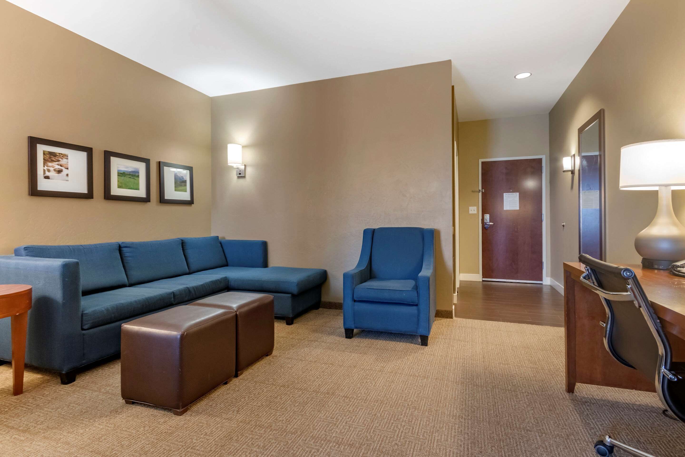 A hotel room with a blue sofa, a matching armchair, a wooden coffee table, and a desk with a lamp.