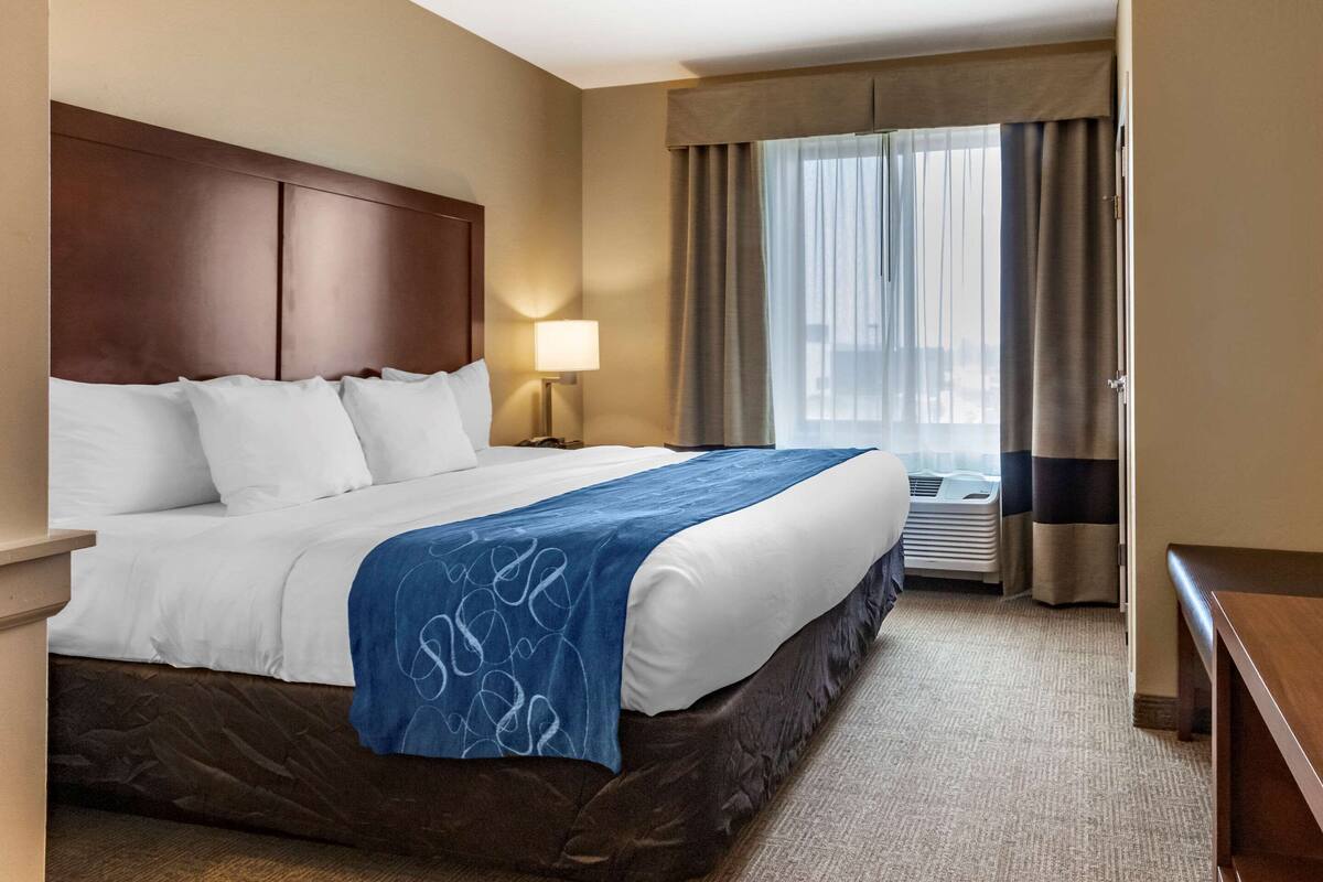 A hotel room with a large bed, a nightstand with a lamp, a window with curtains, and an air conditioning unit.