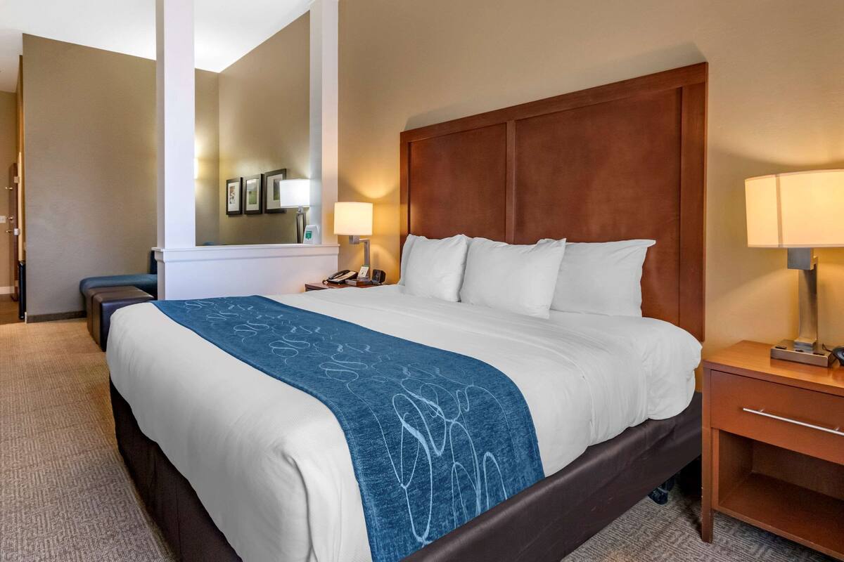 A hotel room with a large bed, a wooden headboard, a bedside table with a lamp, and a mirror on the wall.