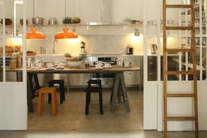 A modern kitchen with white cabinets, a marble countertop, and orange pendant lights. There are stools and a wooden ladder near the kitchen entrance.