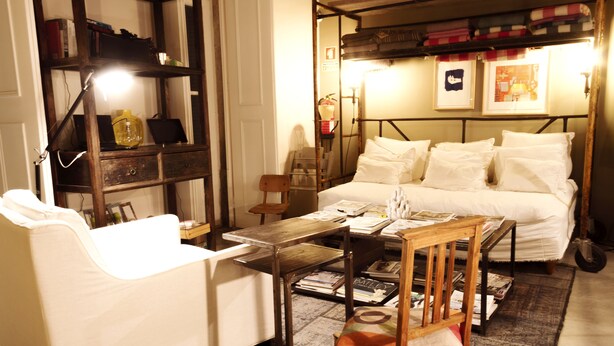 Una habitación acogedora con una cama, un sillón blanco, una mesa de madera y una estantería.