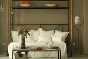 A room with a bed, a table with books and glasses, a vase with flowers, and a shelf with oranges.