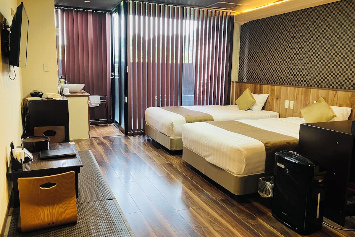 A hotel room with two beds, a desk, a chair, a small table, a trash bin, and a black suitcase.