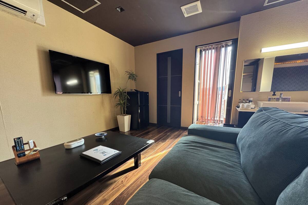 A living room with a flat-screen TV, a sofa, a coffee table, a potted plant, a sliding door, and a bathroom visible through the door.