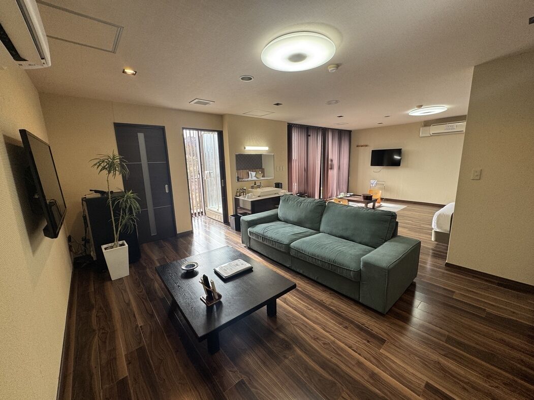 A modern living room with a sofa, a coffee table, and a TV. There is a door leading to a bathroom and a window with curtains.