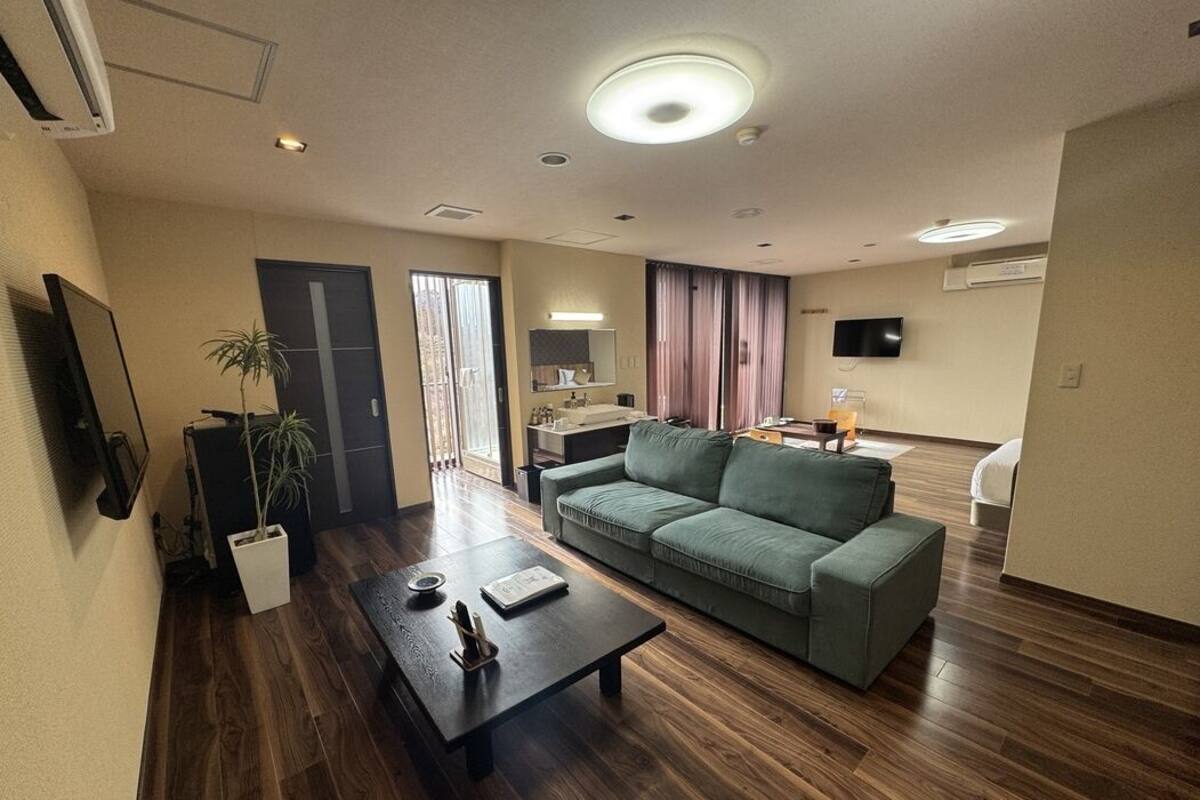 A modern living room with a sofa, a coffee table, and a TV. There is a door leading to a bathroom and a window with curtains.