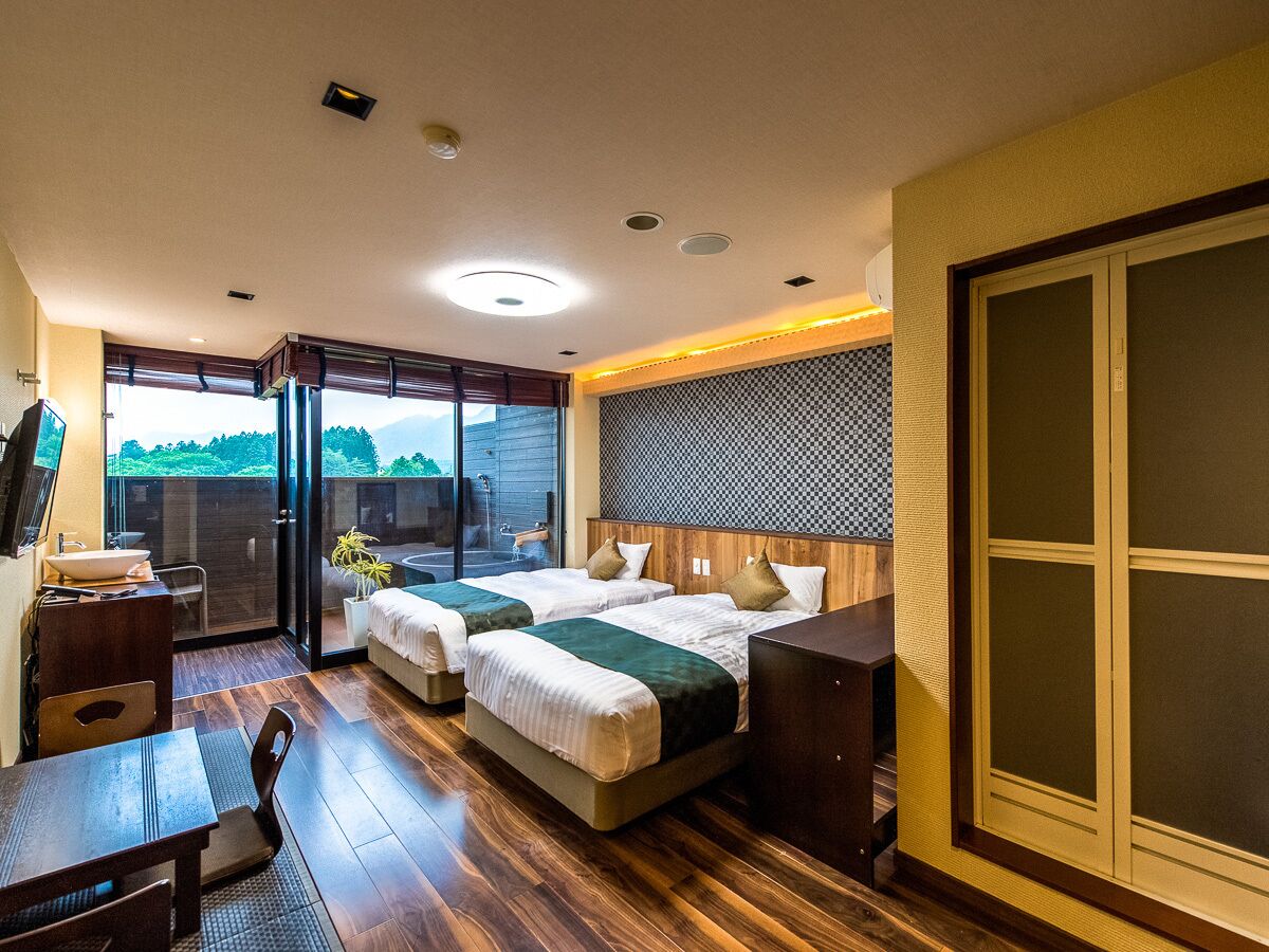 A hotel room with two beds, a desk, a chair, a large window, and a sliding door.