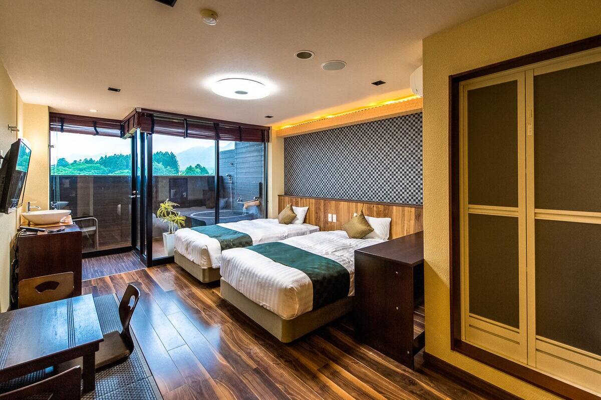 A hotel room with two beds, a desk, a chair, a large window, and a sliding door.