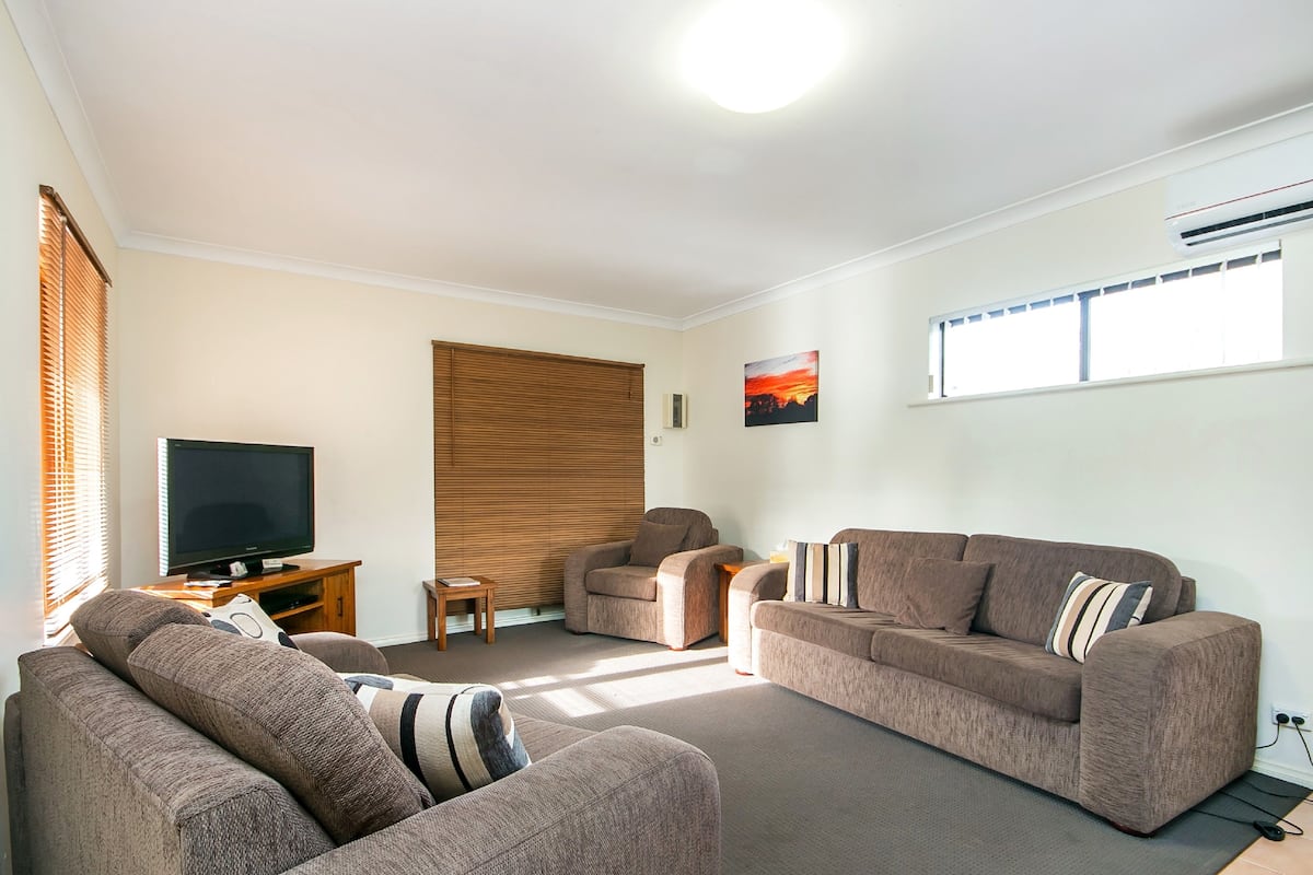 A living room with a flat-screen TV, two sofas, a wooden wall panel, and a window with blinds.