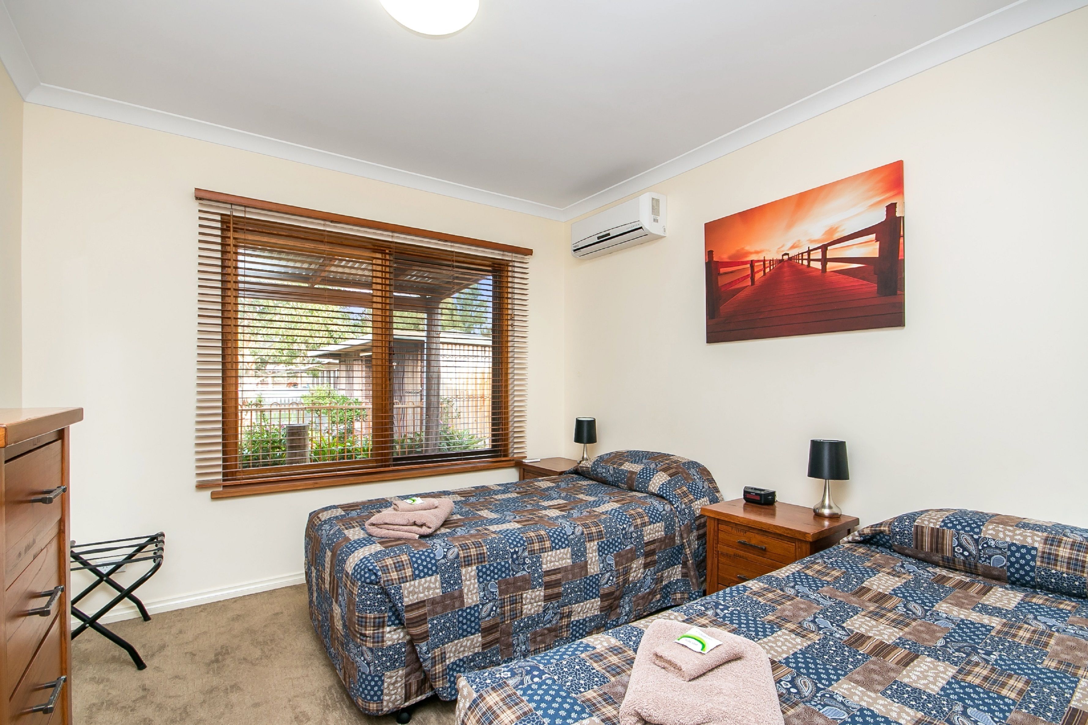 A bedroom with two beds, a window with blinds, an air conditioner, a painting, a desk, and a lamp.