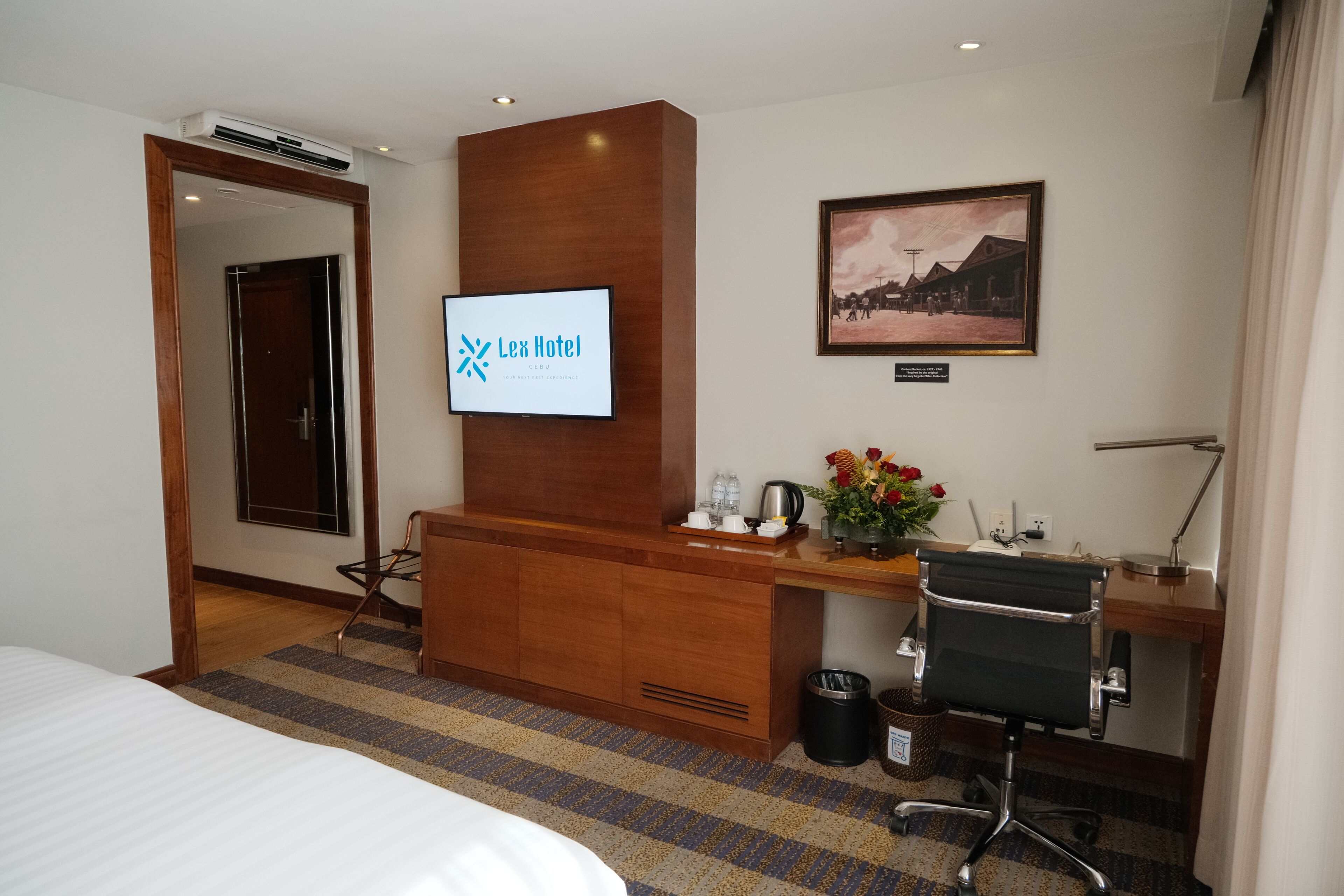 Une chambre d’hôtel avec un lit, un bureau et une chaise, une télévision, un tableau encadré au mur et une poubelle.
