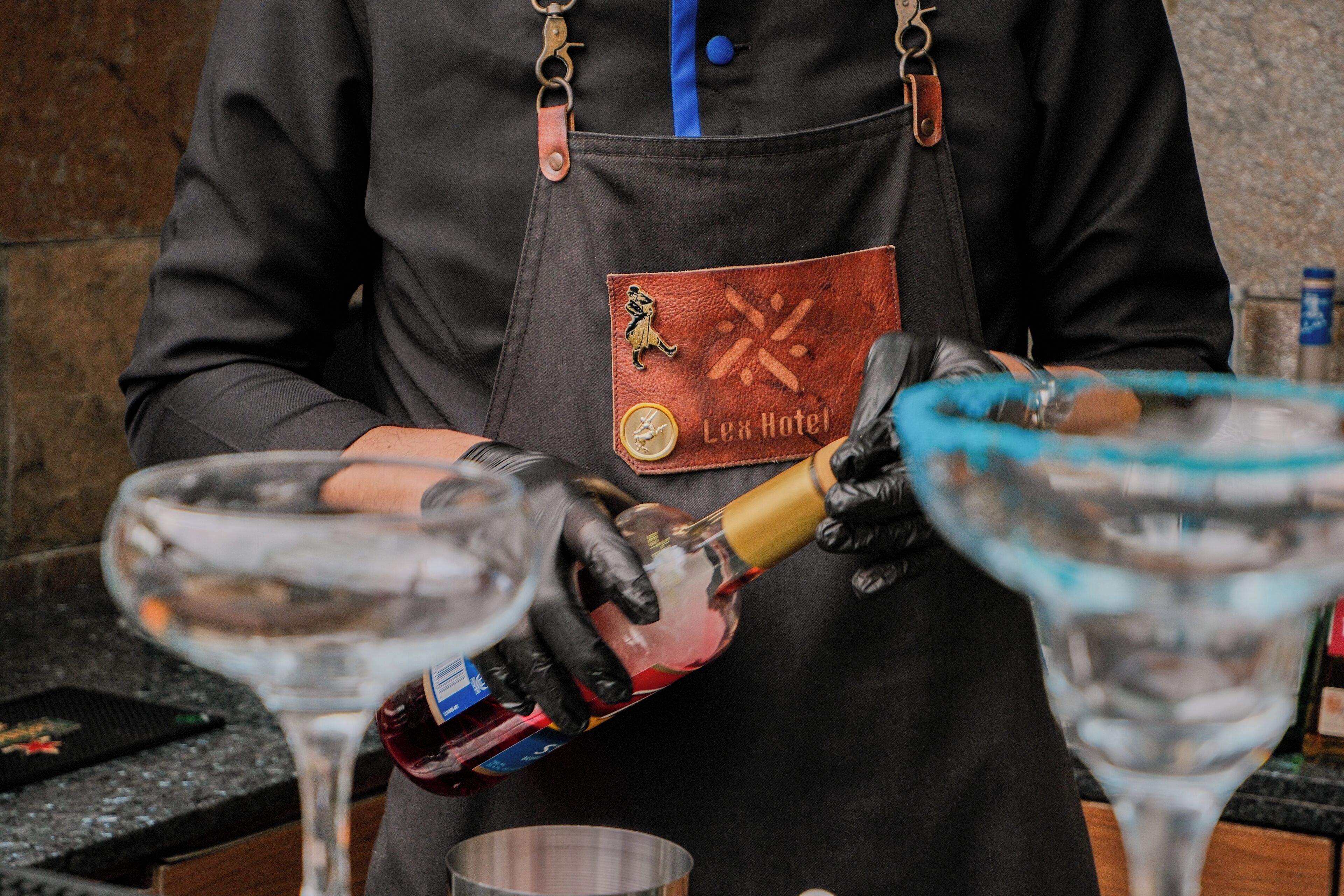 A person in a black apron with a badge, pouring a drink into a martini glass.