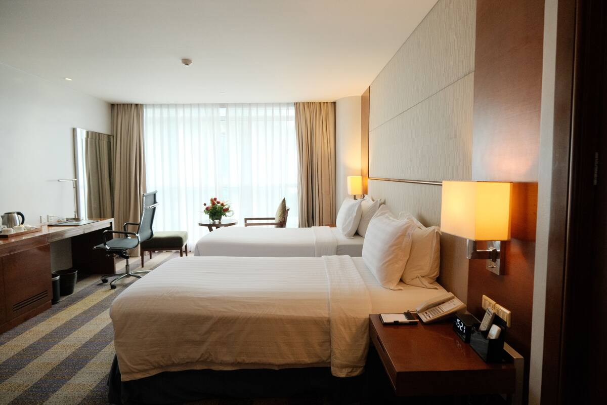 A hotel room with two beds, a desk, a chair, a lamp, and a clock.