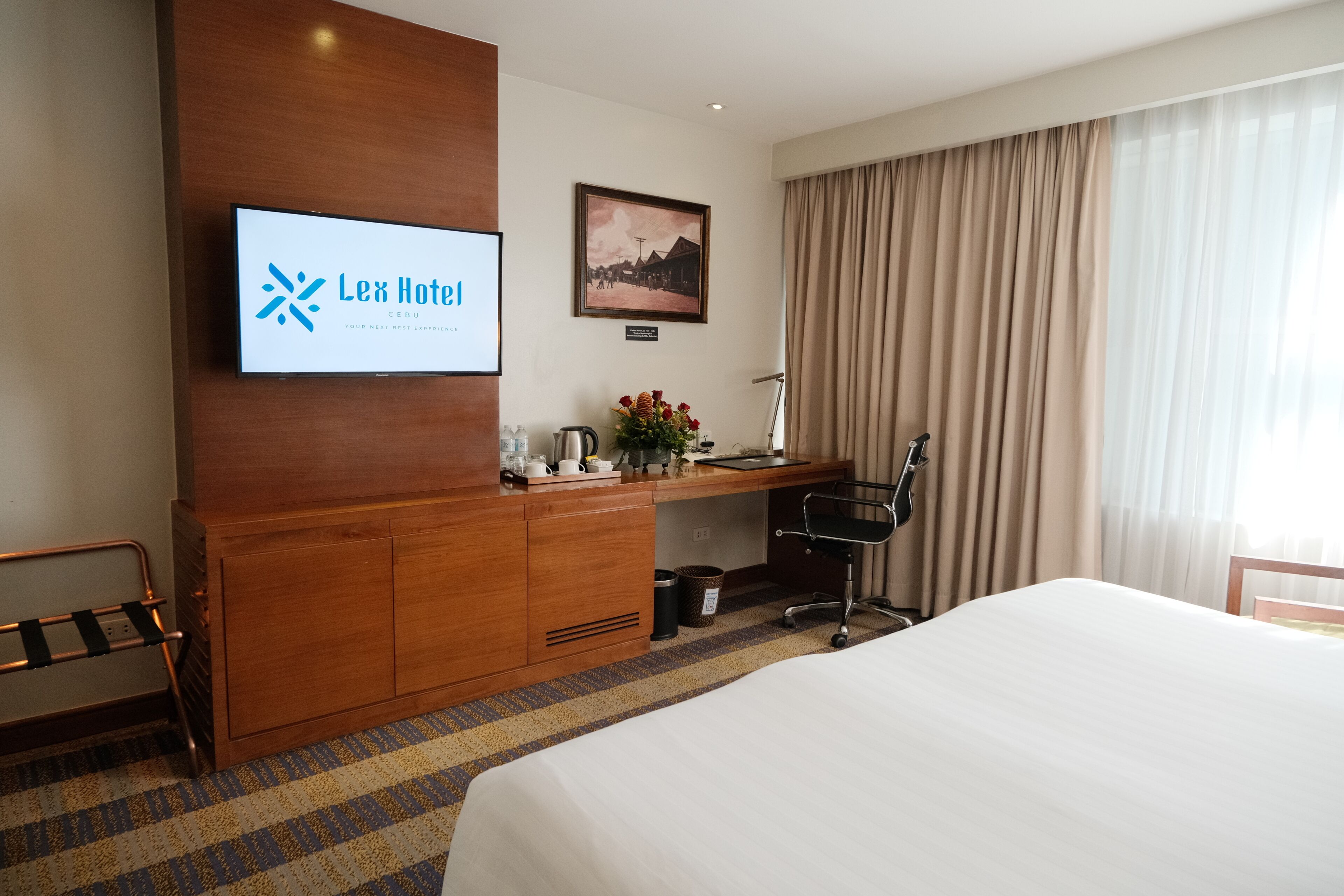 Habitación de hotel con cama, escritorio con silla, televisor mostrando el nombre del hotel, un cuadro en la pared y ventana con cortinas.