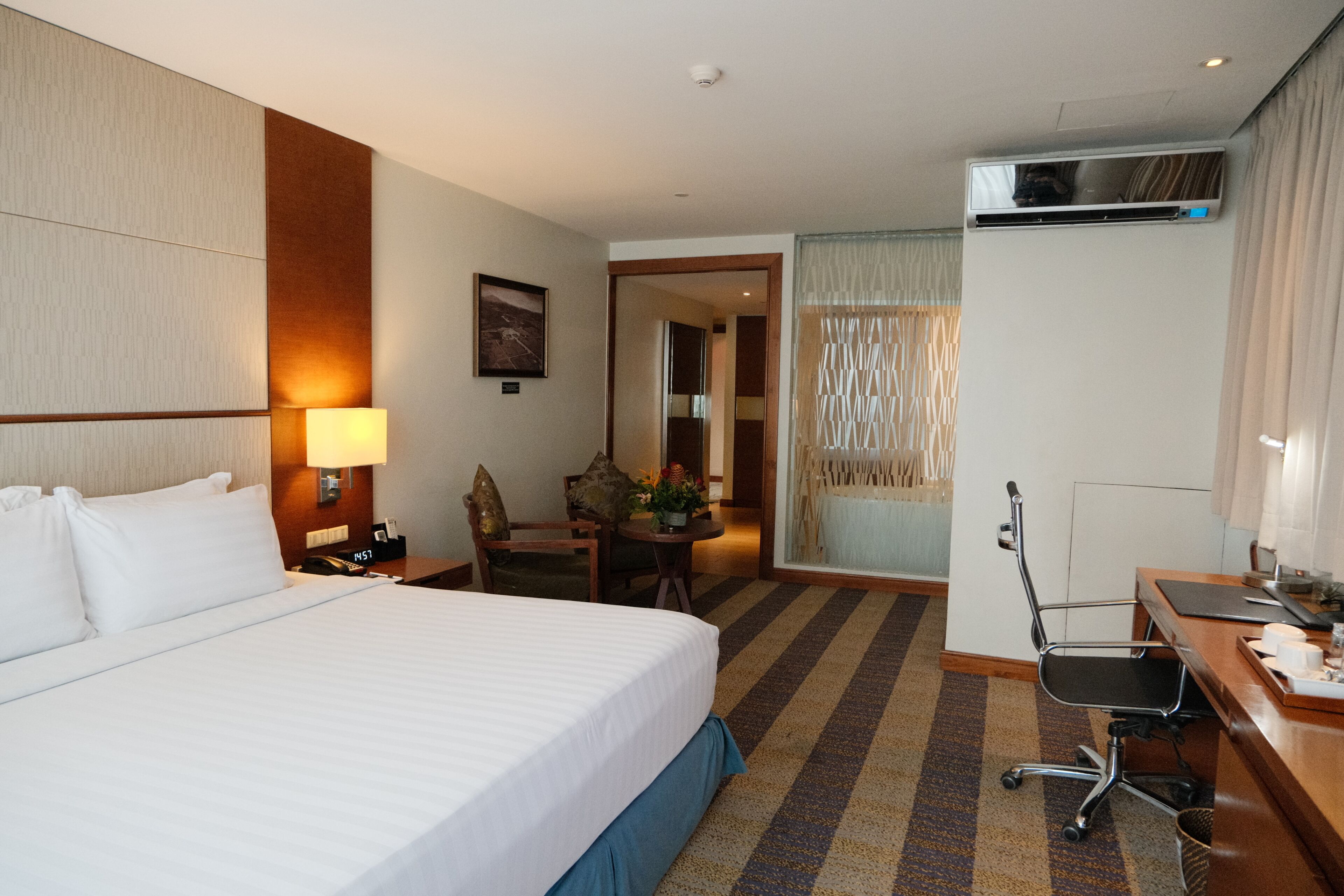 A hotel room with a large bed, a desk with a chair, a small table with a lamp, and a door leading to another room.