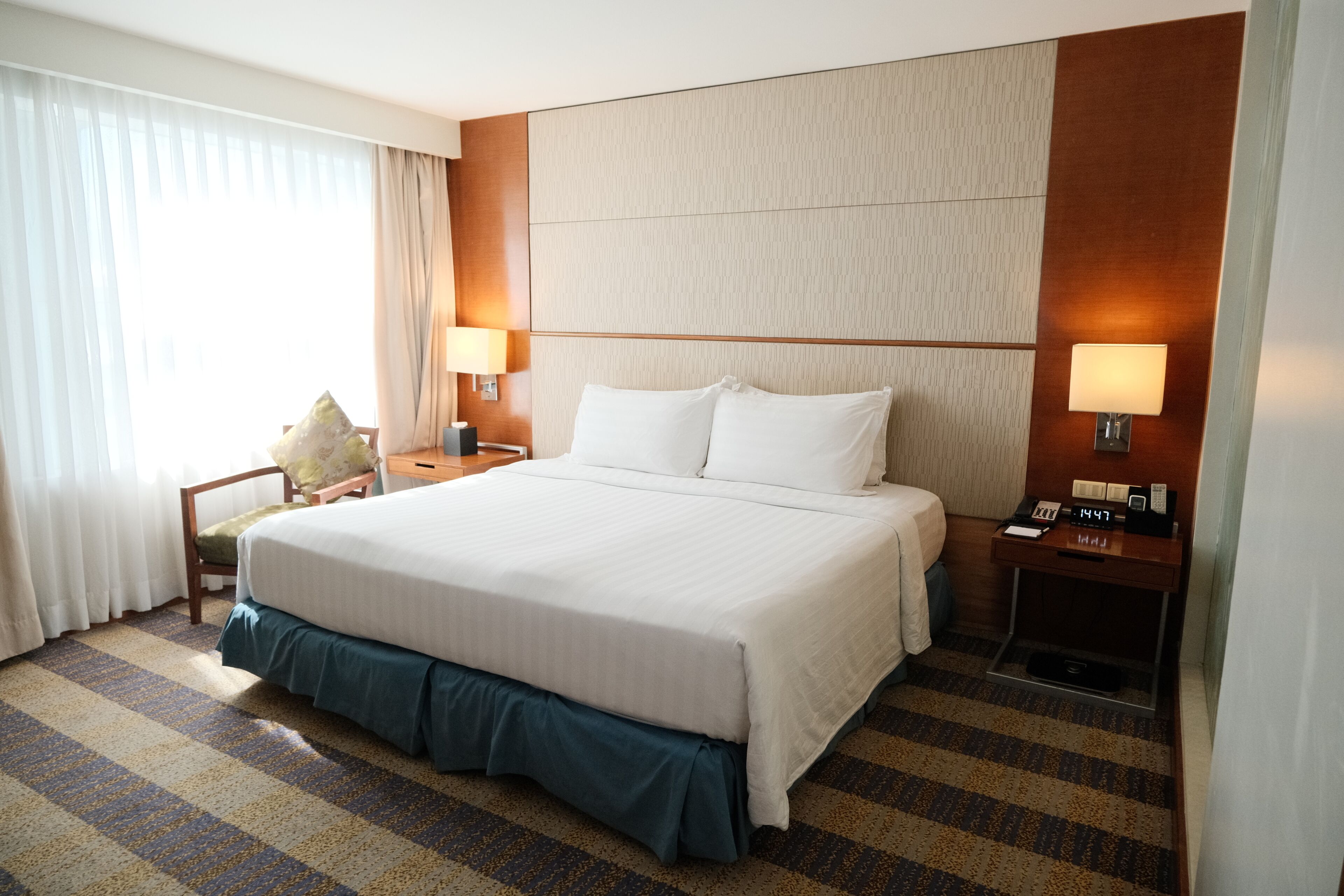 Une chambre d’hôtel avec un grand lit, deux tables de chevet avec des lampes, une chaise et une petite table de nuit avec une horloge.
