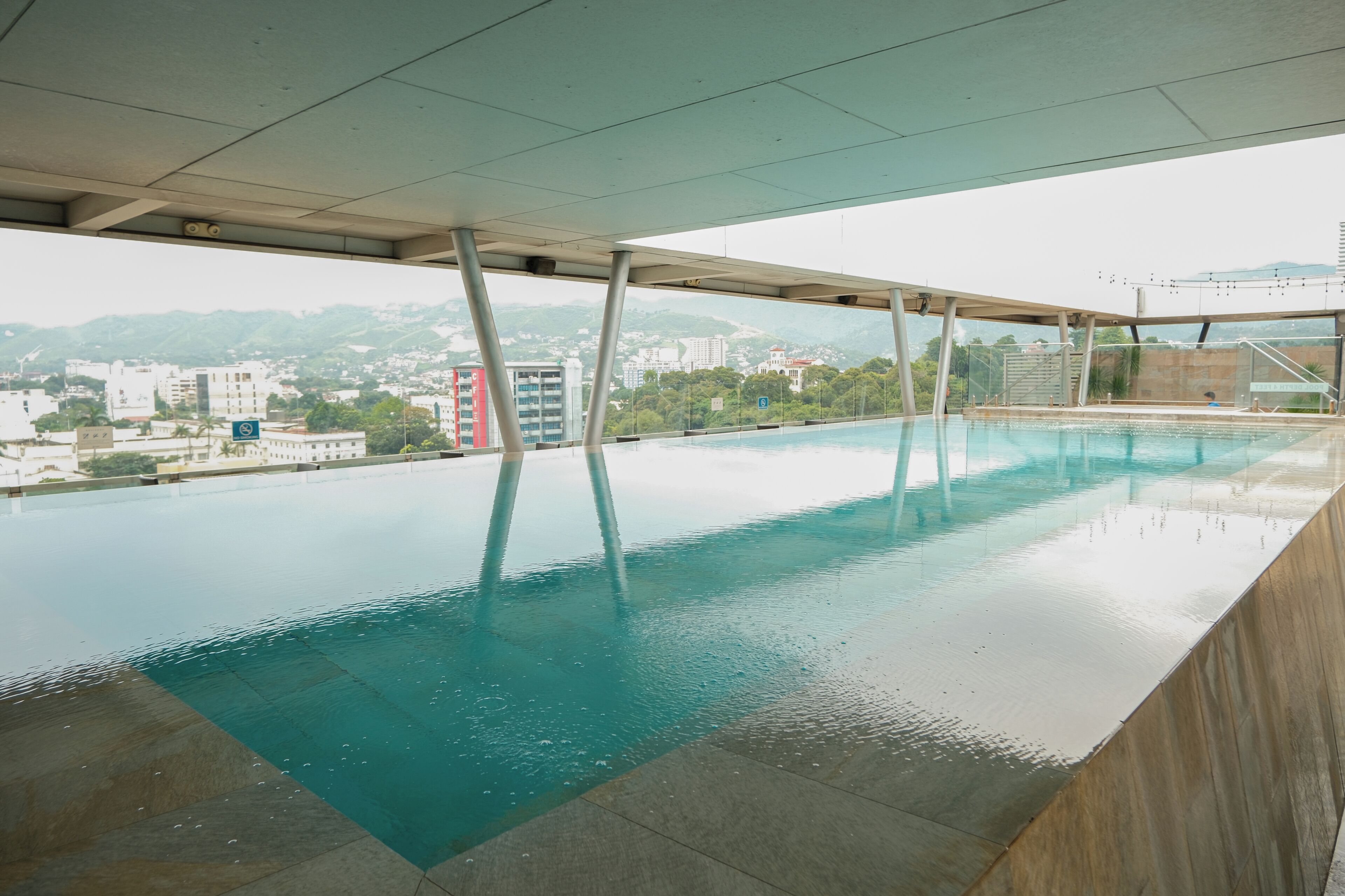 An indoor swimming pool with clear water, a view of buildings and hills in the background, and a modern architectural design.