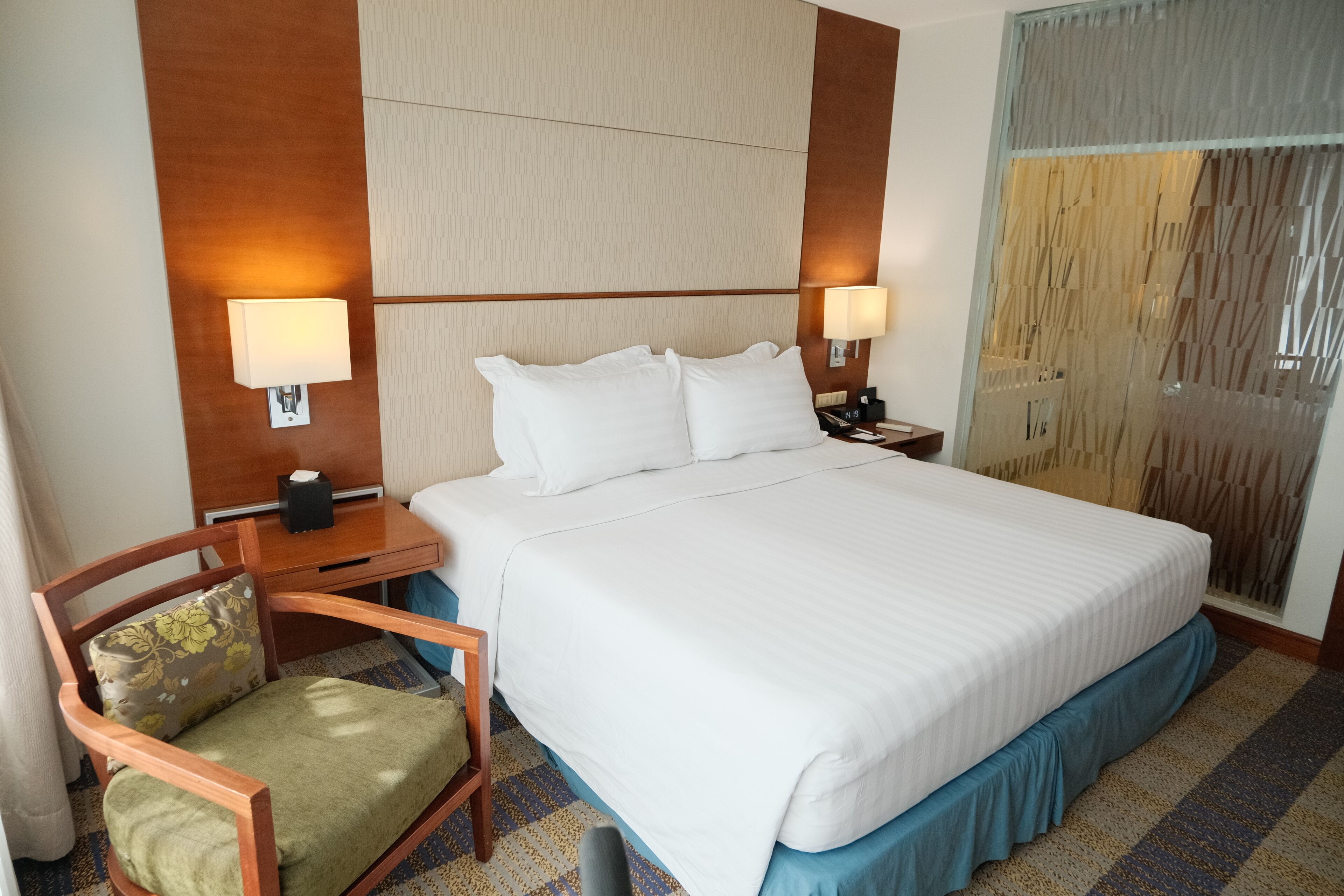 Habitación de hotel con una cama grande, dos lámparas de noche, una mesita de noche de madera, una silla con cojín estampado y una alfombra con diseño.