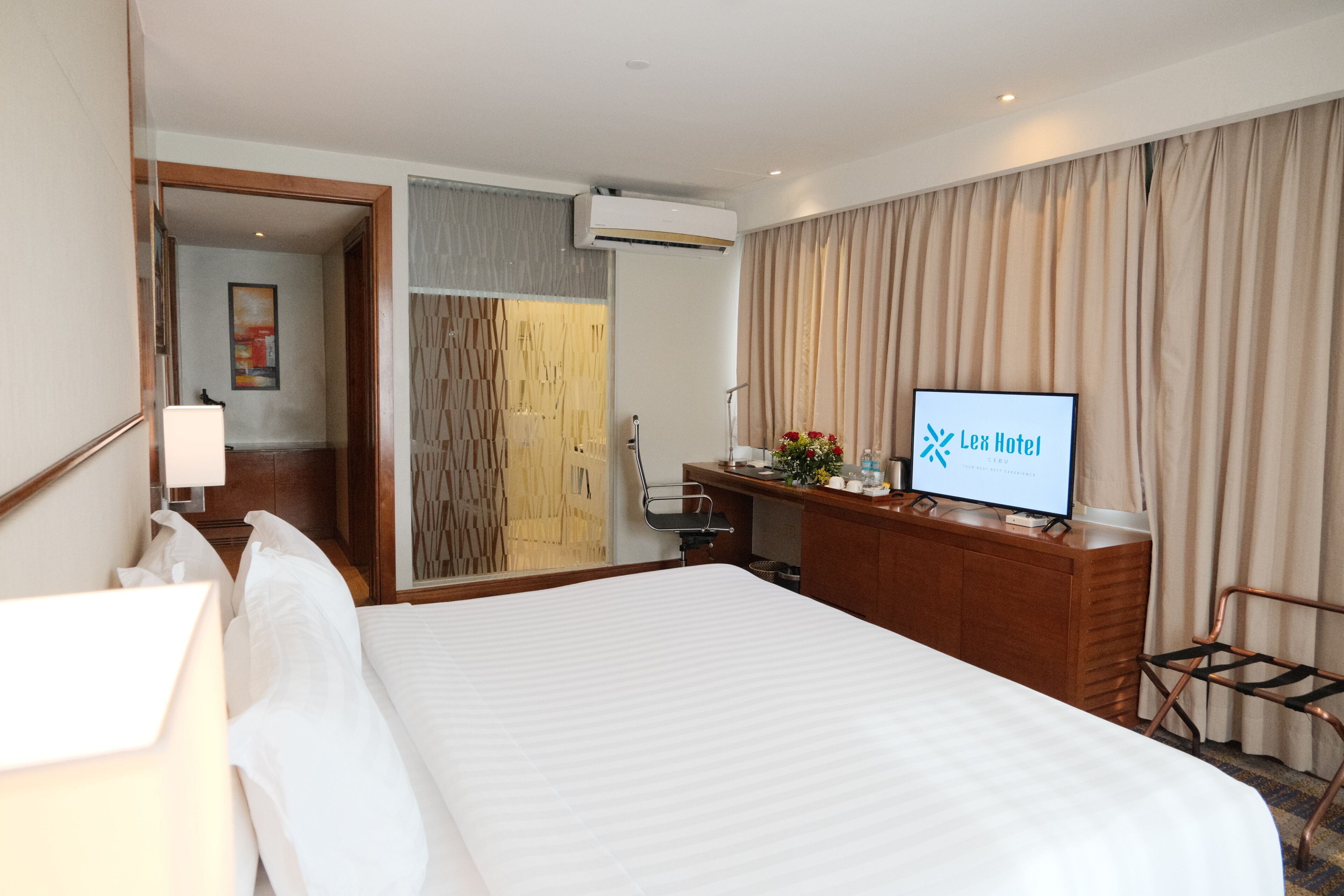 A hotel room with a bed, a desk with a TV, a mirror, and a door leading to another room.