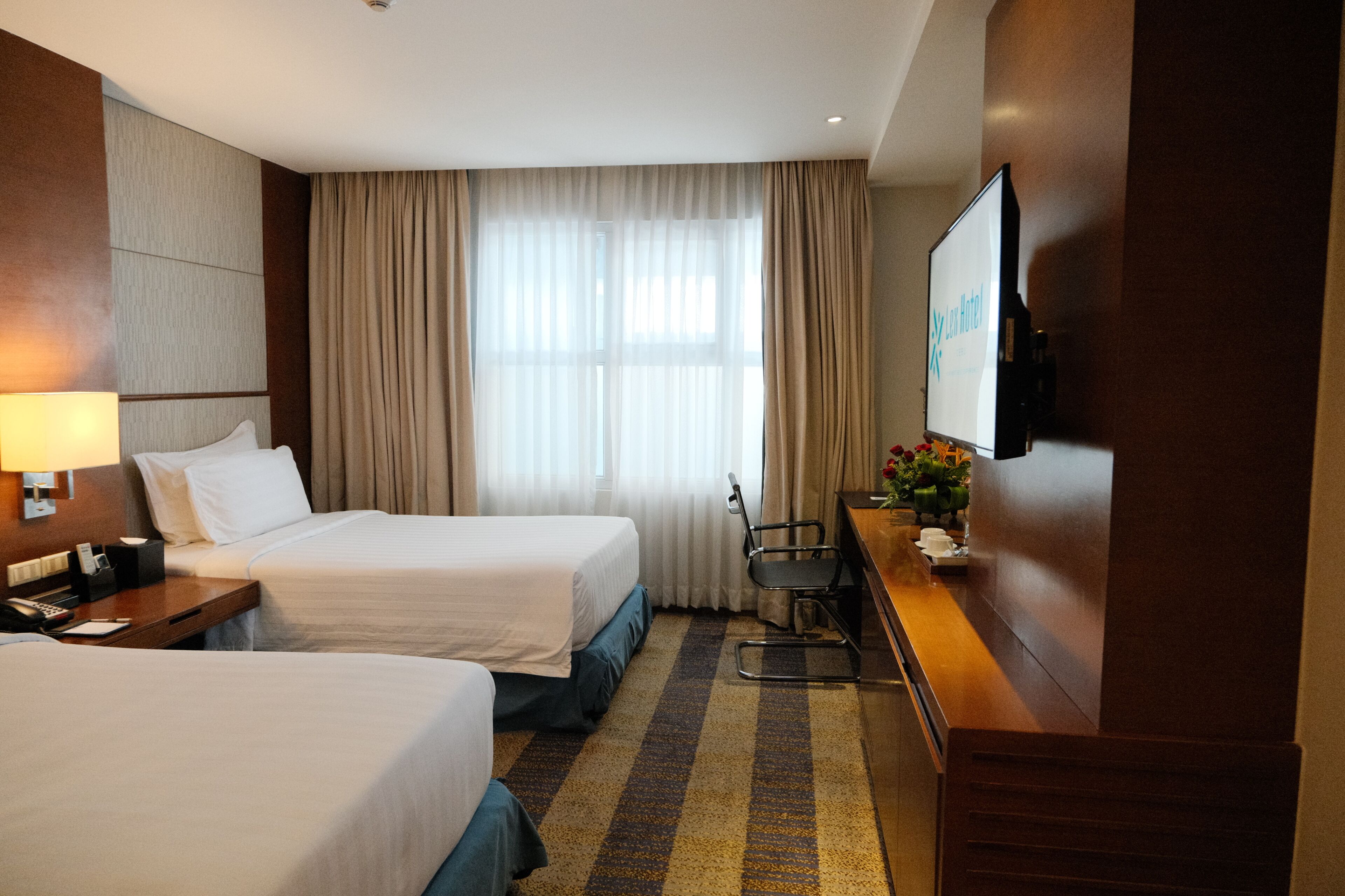 A hotel room with two beds, a desk with a chair, a television, and a window with curtains.