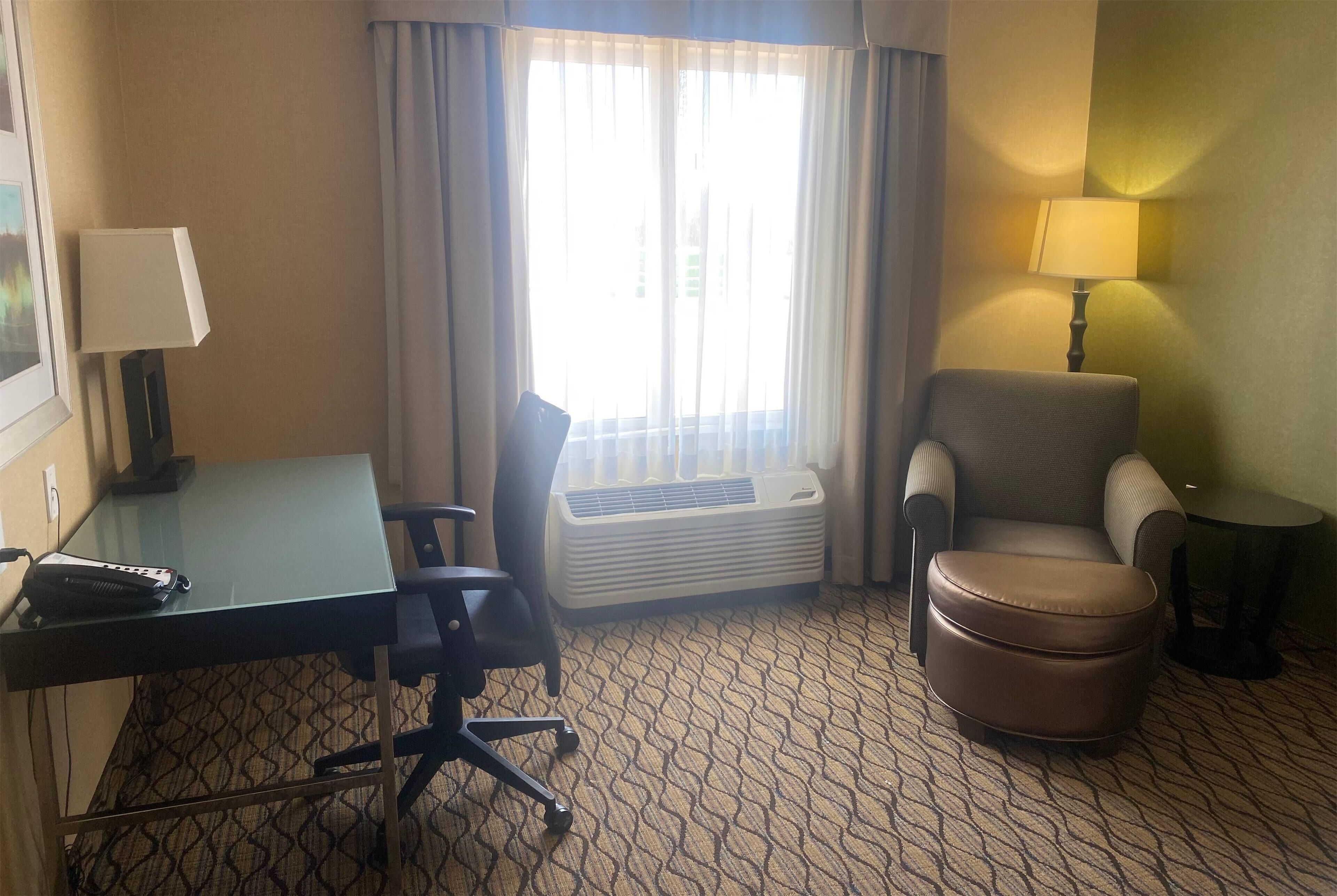 Una habitación de hotel con escritorio, silla de oficina, sillón, otoman y lámpara de pie.