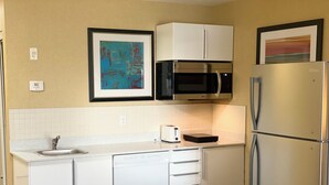 A compact kitchen with white cabinets, a stainless steel refrigerator, a microwave, and a toaster. There is a framed abstract painting on the wall.