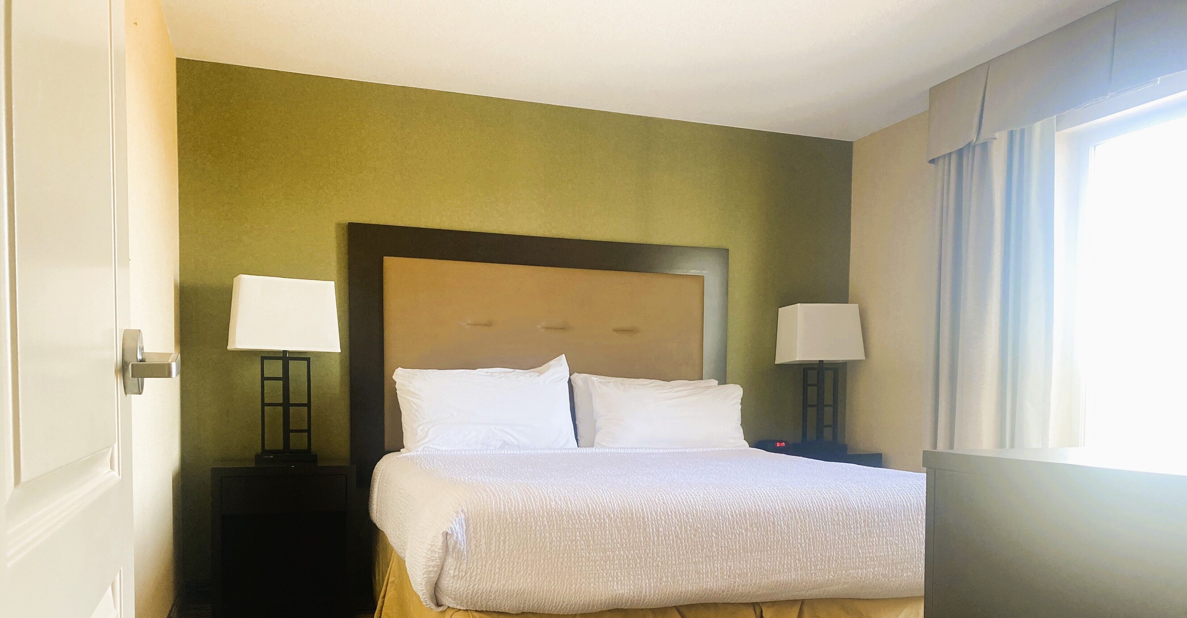 Habitación de hotel con una cama grande, dos mesitas de noche con lámparas, una pared verde y una ventana con cortinas.