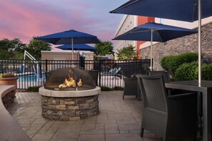 Un patio al aire libre con una chimenea de piedra, mobiliario exterior, sombrillas y una piscina al fondo.