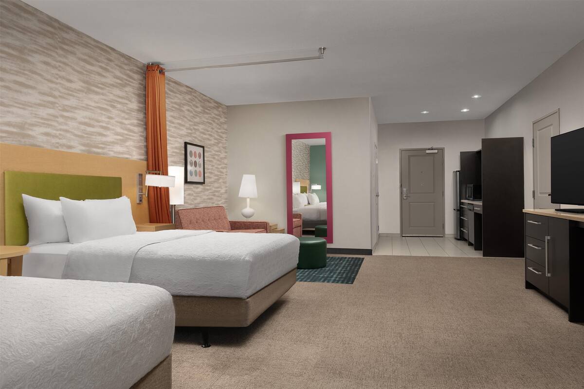 Une chambre d’hôtel avec deux lits, un bureau, un miroir, une télévision et une kitchenette.