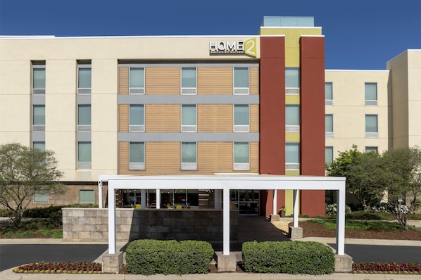 Ein mehrstöckiges Hotel mit moderner Fassade, einer überdachten Eingangsparte und einer angelegten Grünfläche mit Sträuchern und Blumen.
