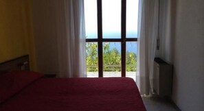 A bedroom with a bed, a window with curtains, and a view of greenery outside. - Hotel Due Gemelli (Riomaggiore)