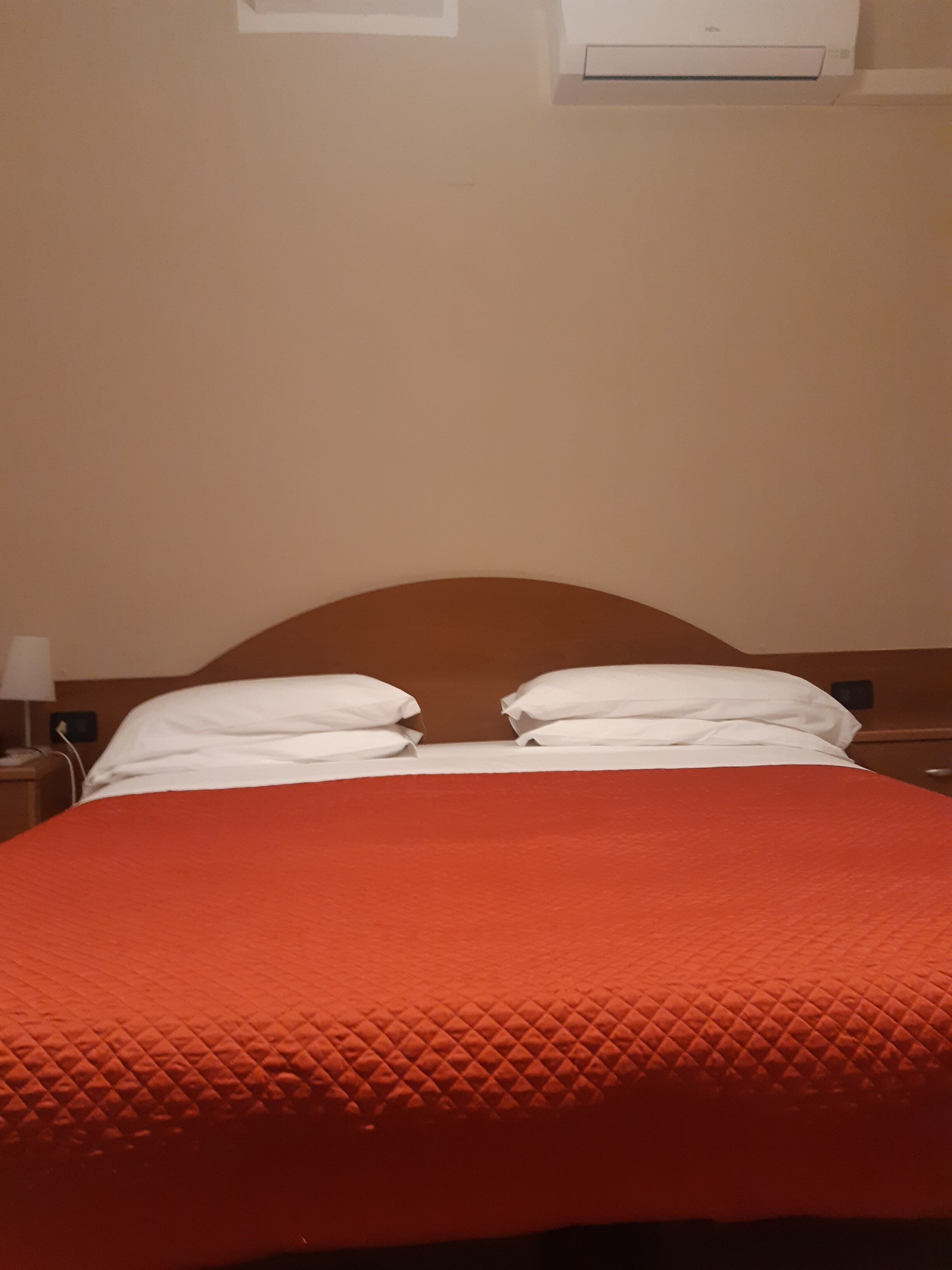 Un lit recouvert d’une courtepointe orange à motifs et agrémenté d’oreillers blancs, une tête de lit en bois et une table de chevet avec une lampe.