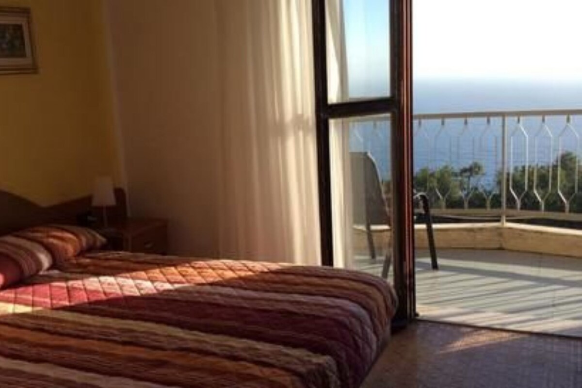 A hotel room with a bed, a nightstand, a window with a view, and a balcony with a railing.