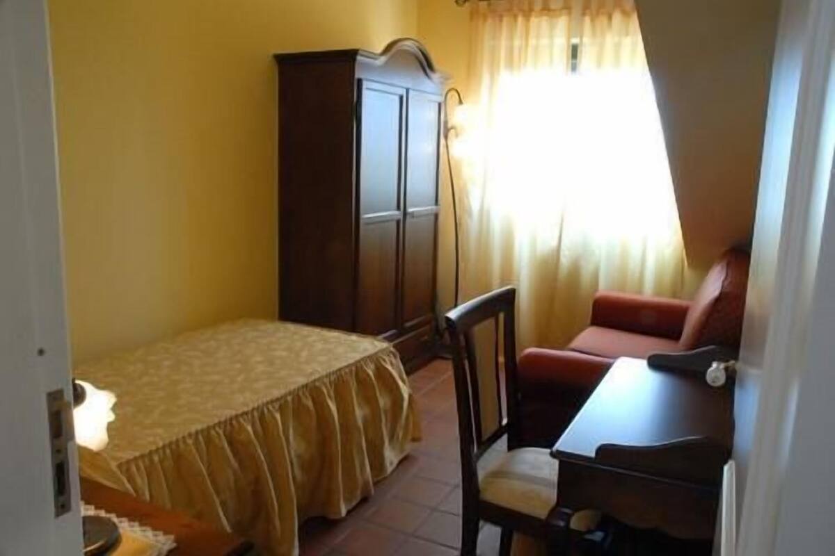 A bedroom with a bed, a wooden wardrobe, a desk with a chair, and a window with curtains.