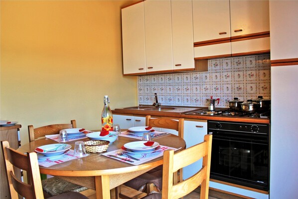 A dining area with a wooden table set for four, surrounded by wooden chairs. A kitchen with white cabinets and a tiled backsplash is visible in the background.