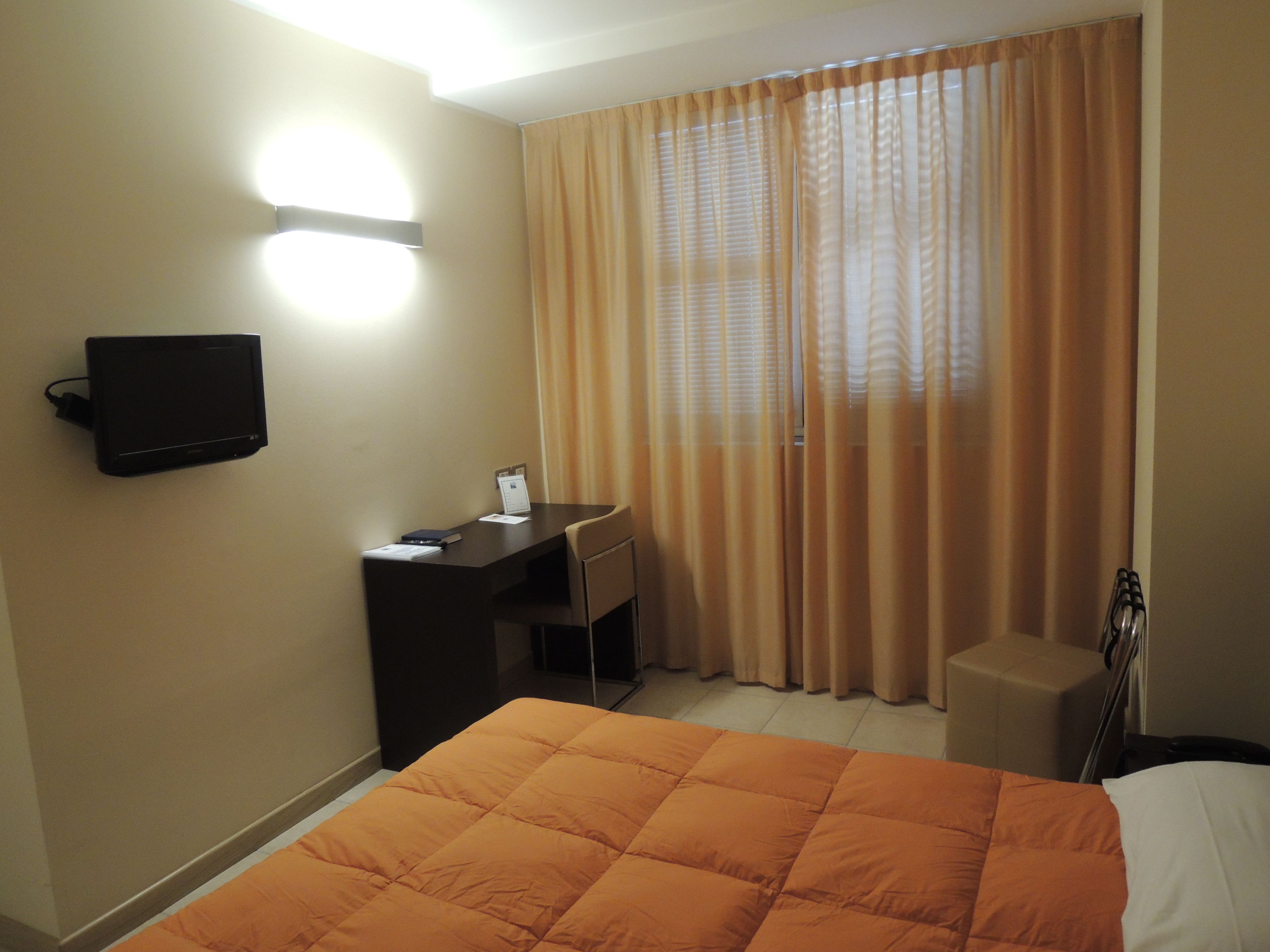 A hotel room with a bed, a desk, a chair, a television mounted on the wall, and a window with curtains.