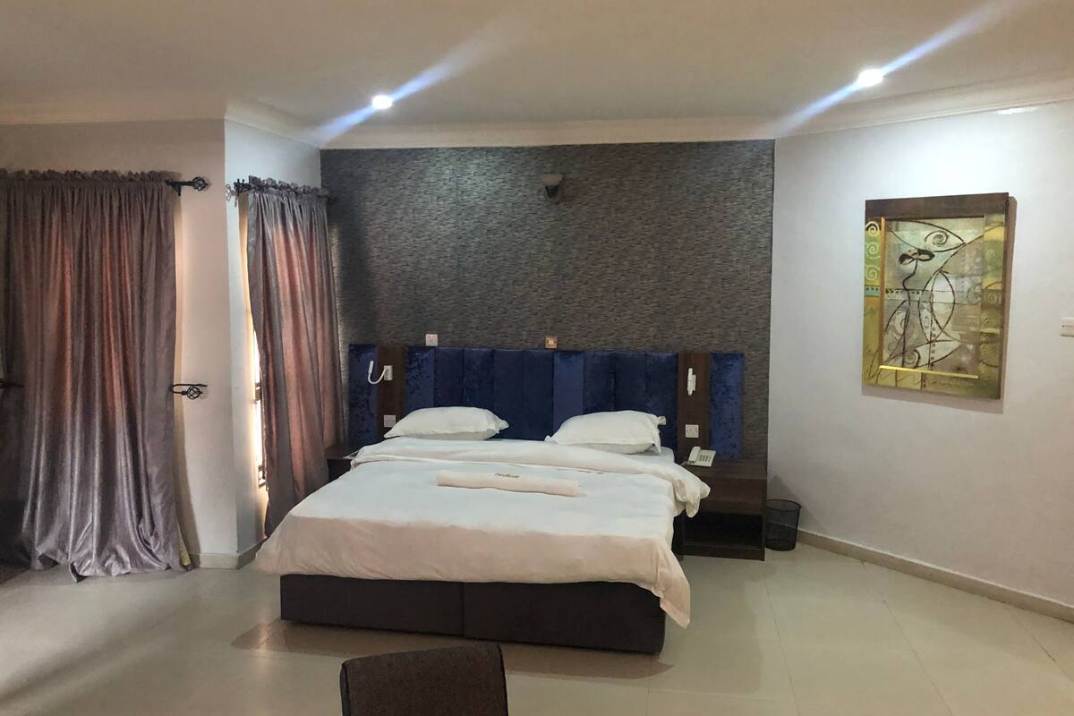 A modern hotel room with a large bed, a headboard with a textured wall, a bedside table, a telephone, and a painting on the wall.