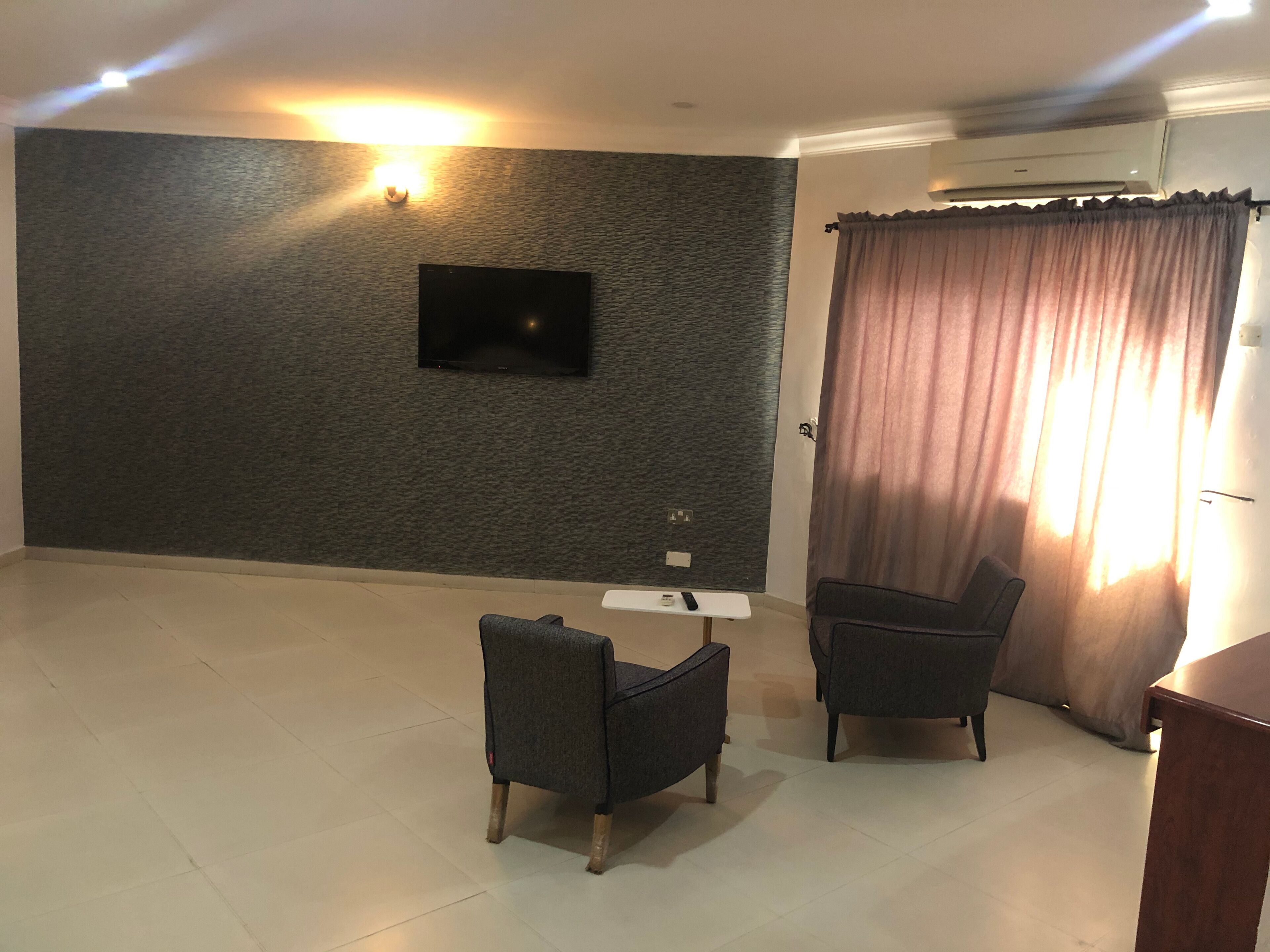 A modern living room with a flat-screen TV mounted on a textured wall, two armchairs, a small white table, and a large window with curtains.