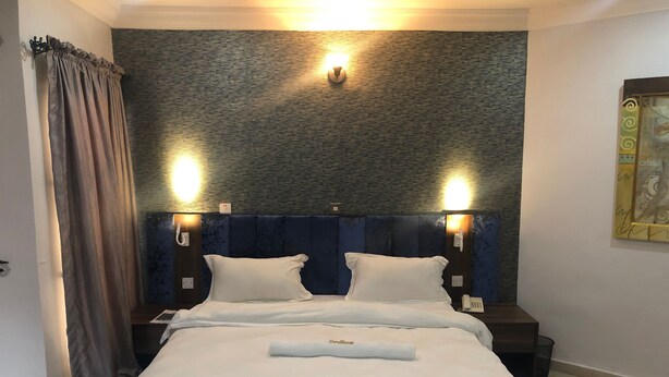 A hotel room with a bed, two bedside tables, a wall-mounted telephone, and a painting on the wall. - Solitude Hotel Victoria Island (Lagos)