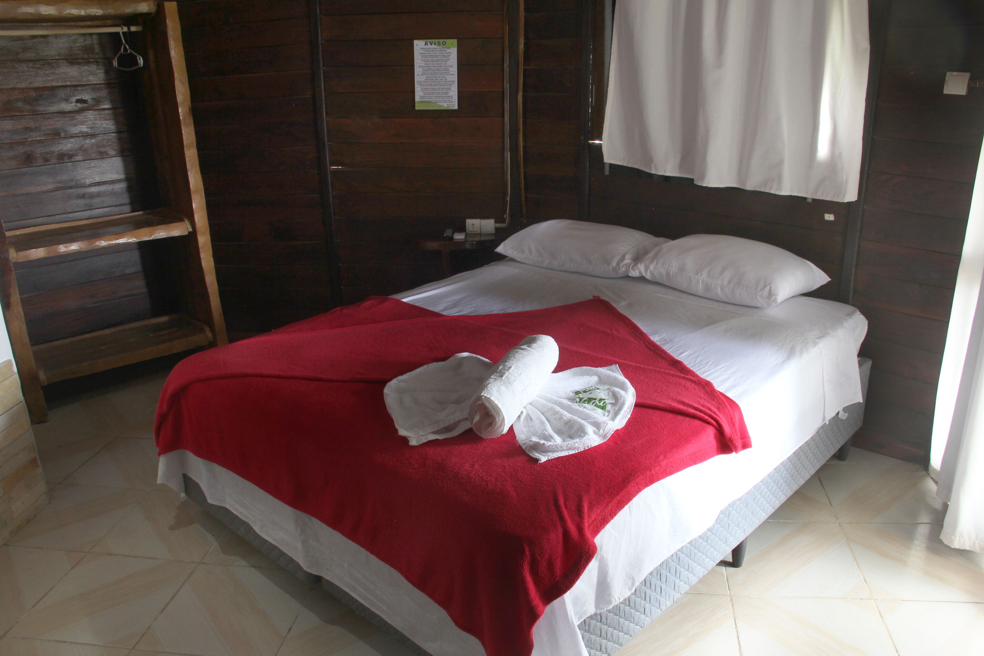 Ein Einzelbett mit einer rot-weißen Tagesdecke, einem weißen Kopfkissen und einem aufgerollten weißen Handtuch.