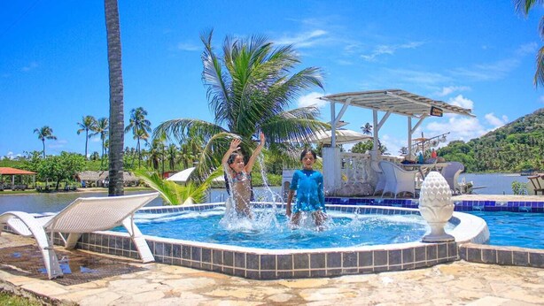 A swimming pool with two people playing, surrounded by palm trees and a water slide. - Pousada Paradiso Tropical (Marechal Deodoro)