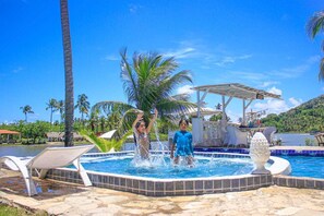 A swimming pool with two people playing, surrounded by palm trees and a water slide. - Pousada Paradiso Tropical (Marechal Deodoro)