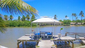 A wooden dock with a canopy and seating area overlooking a calm body of water, surrounded by lush greenery and palm trees. - Pousada Paradiso Tropical (Marechal Deodoro)