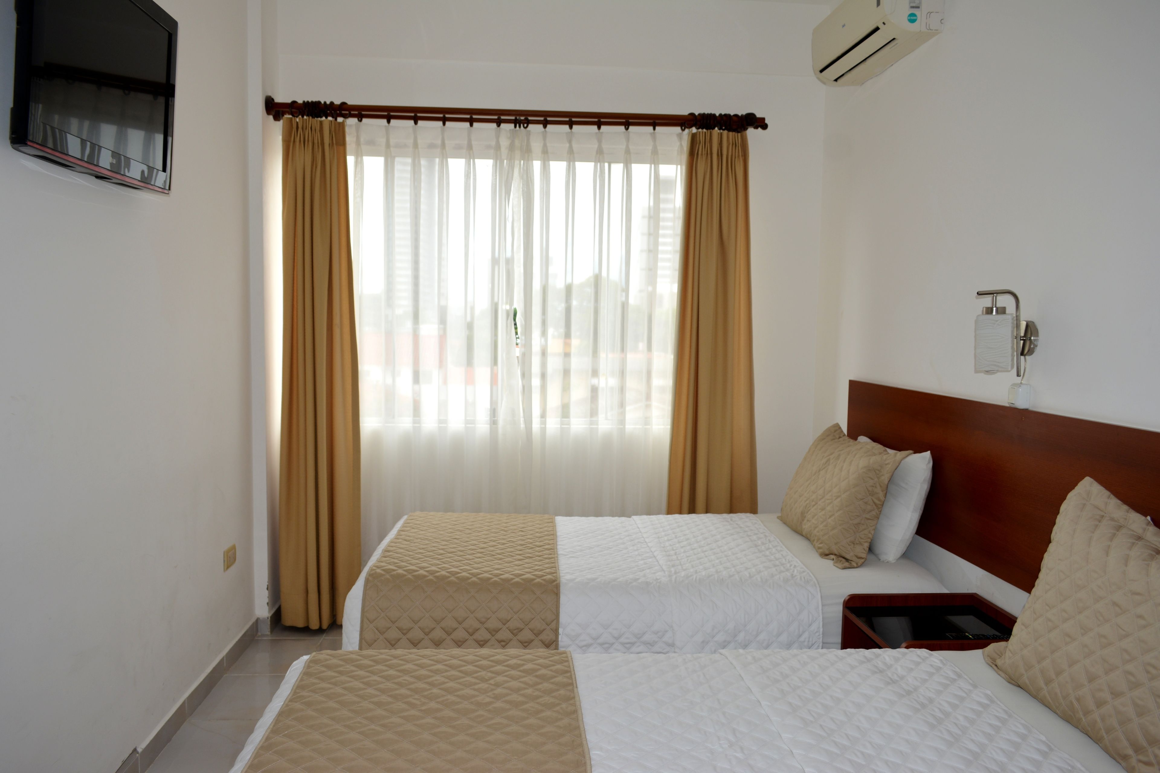 A hotel room with two beds, beige and white bedding, a wooden headboard, a bedside table with a tray, and a window with sheer curtains.