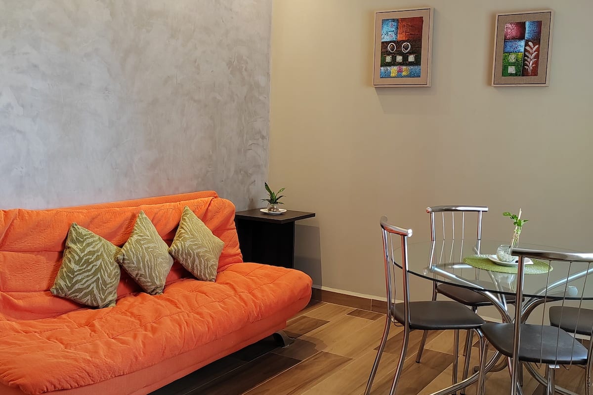 A modern living room with a sofa, a glass dining table, and metal chairs.